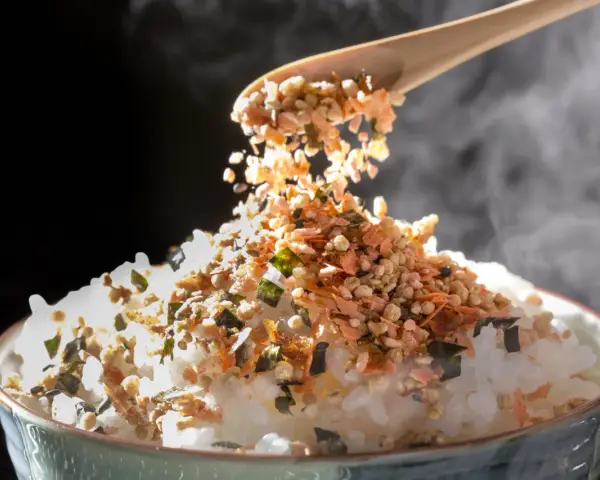 Łyżka sypie na parujący ryż kolorowy furikake. To japońska posypka, która dodaje smaku.