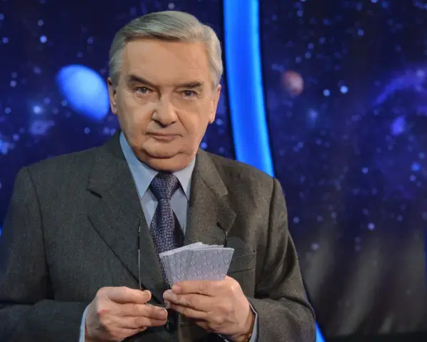Starsza osoba w garniturze trzyma karty z napisem "1 z 10", gotowa do emisji.