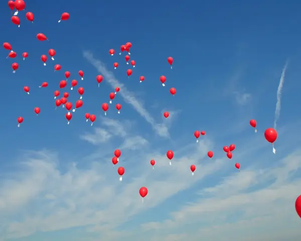 Många röda heliumballonger stiger mot en klarblå himmel med tunna moln.