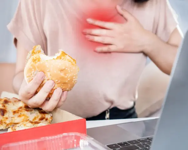 Kobieta z bólem w klatce piersiowej trzyma hamburgera. Zastanawia się, co pić na zgagę po zjedzeniu pizzy i fast foodów.