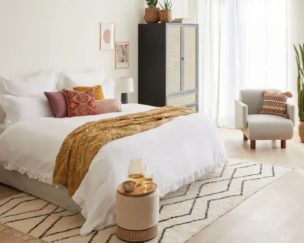Habitación serena con cama blanca, manta ocre y cojines coloridos. Un armario de ratán y una silla acogedora complementan el estilo feng shui cama.