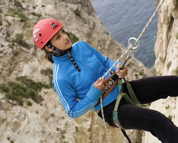 Junge Frau beim Abseilen an einer Felswand, Malta Klettern. Sie trägt einen roten Helm und eine blaue Jacke.