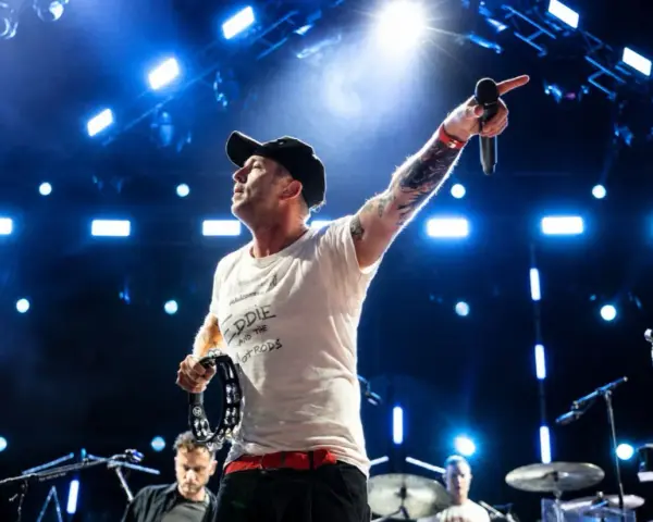 Wokalista z tatuażami i czapką na głowie prowadzi koncert One Republic, wskazując ręką w górę i trzymając tamburyn.