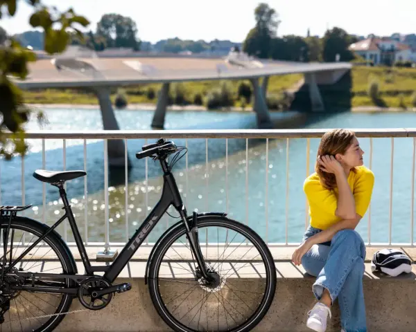 En kvinna i gula tröjan och jeans vilar vid en elcykel. Frågan "hur lång tid tar det att cykla 1 mil med elcykel" hänger i luften.