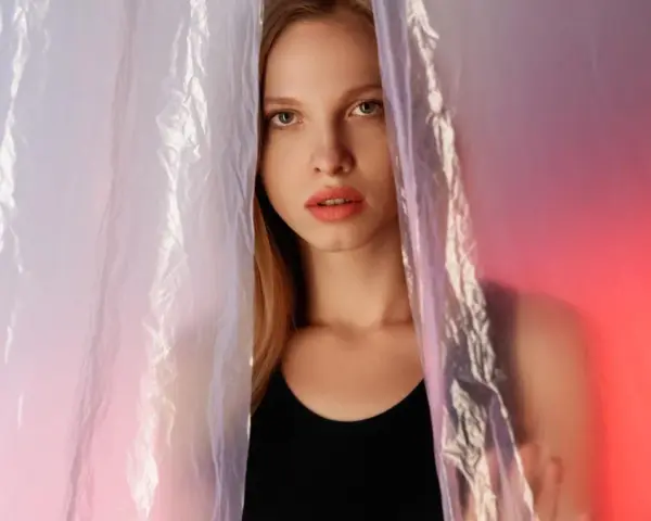 A young woman peeks through crinkled plastic sheet materials, her face illuminated by soft, colorful light.