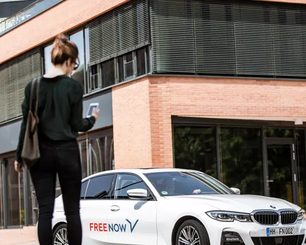 Frau mit Smartphone wartet auf FREE NOW Fahrt. Keine Beschwerde, nur Vorfreude auf die Fahrt.