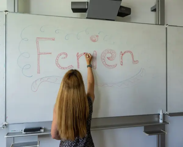 Frau schreibt "Ferien" an Tafel, Vorfreude auf Pfingstferien Rheinland-Pfalz.
