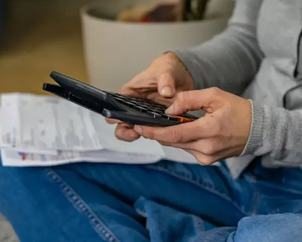 Ręce osoby w dżinsach i szarej bluzie liczą coś na kalkulatorze. Obok leżą dokumenty. Może to być rozliczenie rachunków, które zajmuje jej g12w godziny.