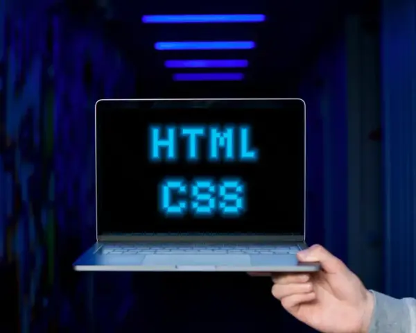 <p>Dłoń trzyma laptopa, na ekranie którego świeci napis "HTML CSS" w niebieskich pikselach. Tło jest ciemne, z niebieskimi neonami.</p>
