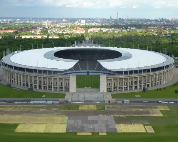 Das Olympiastadion Berlin mit seinem markanten Dach und der Skyline im Hintergrund. In der Ferne ragt der Fernsehturm, der wie ein glockenturm Berlin überragt.