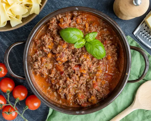 Herzhaftes Ragout Bolognese in einem Topf, garniert mit Basilikum. Daneben frische Tomaten, Bandnudeln und Parmesan.