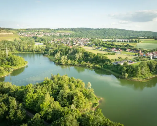 Grüne Hügel, ein Dorf und der glitzernde Klostersee Triefenstein unter blauem Himmel.
