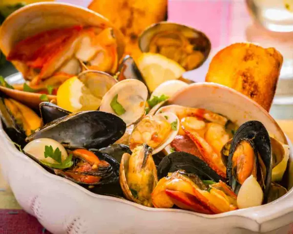 Un fumetto di pesce ricco di cozze, vongole e crostacei, guarnito con limone e prezzemolo, servito in una ciotola bianca.