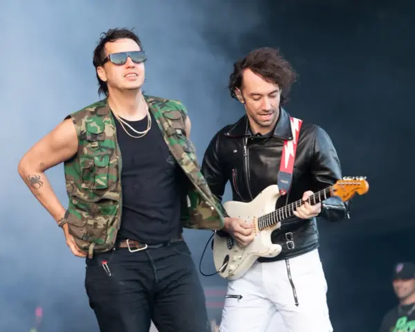 Albert Hammond Jr. w kamizelce moro i okularach stoi obok gitarzysty w skórzanej kurtce, grającego na białej gitarze.