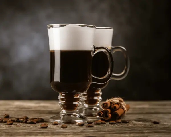 Två glas med irish coffee, toppade med grädde, står på ett träbord. Kaffebönor och kanelstänger ligger utspridda.