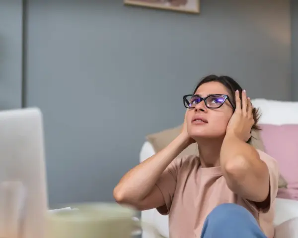 Kobieta z okularami zakrywa uszy, cierpiąc z powodu nadwrażliwości na dźwięki. W tle laptop i kanapa.