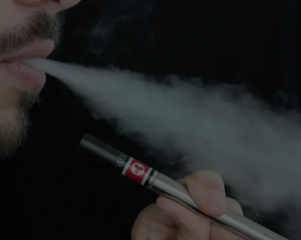 Un hombre con barba exhala vapor de un vapeador, similar al estilo Heisenberg.