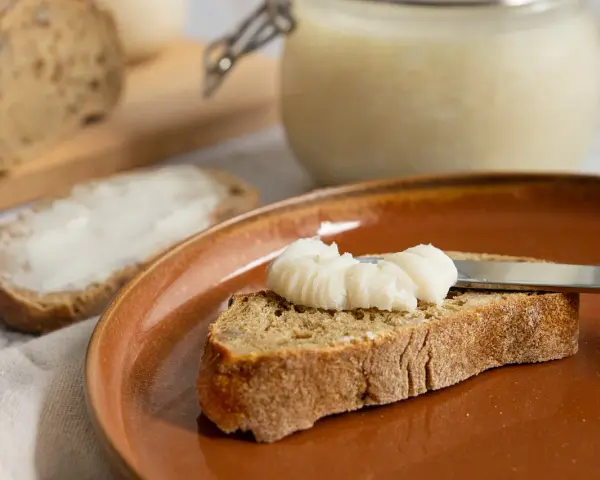 Ein Glas mit selbstgemachtem Schmalz steht bereit. Eine Scheibe Brot wird gerade mit dem Schmalz bestrichen, ein Genuss für jeden, der Schmalz selber machen möchte.