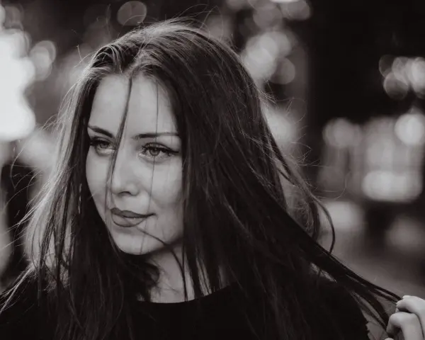 Portret młodej kobiety z burzą ciemnych włosów. Jej spojrzenie jest lekko zadumane, a usta subtelnie uśmiechnięte. Grzywka dodaje jej tajemniczości.
