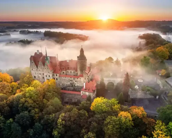 Zamek Książ otoczony jesiennymi drzewami, wyłaniający się z porannej mgły. Słońce wschodzi nad wzgórzami, malując niebo na pomarańczowo.