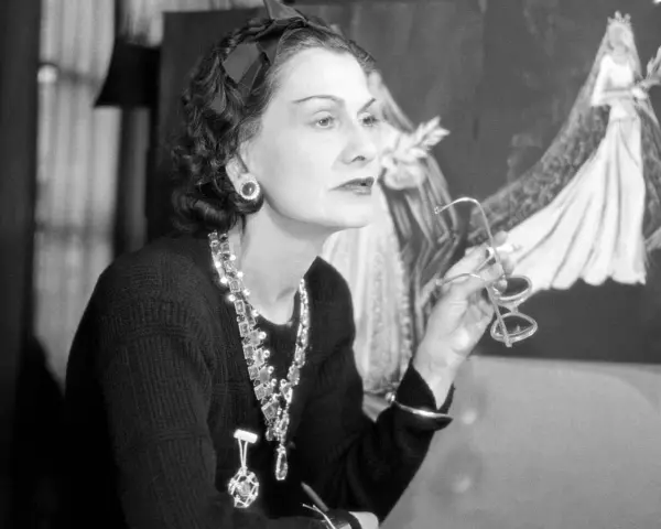 Coco Chanel, icona di stile, con occhiali in mano, sullo sfondo schizzi di abiti da sposa. Un'immagine che racconta la sua storia.