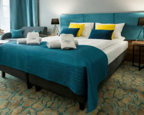 Przytulny pokój hotelowy z dużym łóżkiem, ozdobionym turkusową narzutą i żółto-niebieskimi poduszkami. Ręczniki z logo Stella Resort & Spa zachęcają do relaksu.