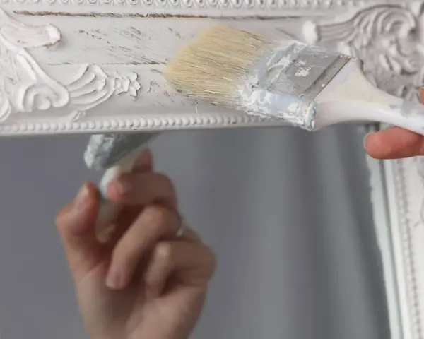 Pintura a la tiza blanco antiguo en un marco de espejo ornamentado. Una mano aplica la pintura con una brocha, creando un acabado desgastado.
