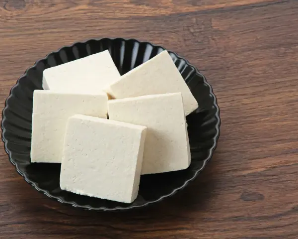 Kilka kostek białego tofu na czarnym talerzu. Czy tofu można mrozić? Tak, po rozmrożeniu zmienia teksturę, stając się bardziej gąbczaste.