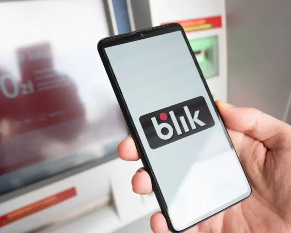 Dłoń trzyma smartfon z logo BLIK przed bankomatem.