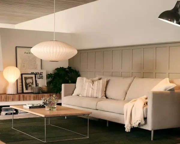 En ljus soffa med kuddar och en filt, framför ett soffbord i trä. En golvlampa och en taklampa lyser upp rummet. Perfekt för att mäta soffa mått.