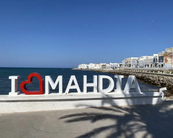 Napis "I ❤️ MAHDIA" na tle morza i białych budynków. Słońce i cień palm. Tunezja Mahdia.