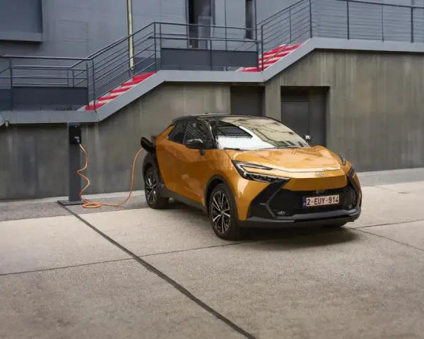 Złoty Toyota plug-in z pomarańczowym kablem ładowania podłączonym do stacji. Nowoczesna architektura w tle.