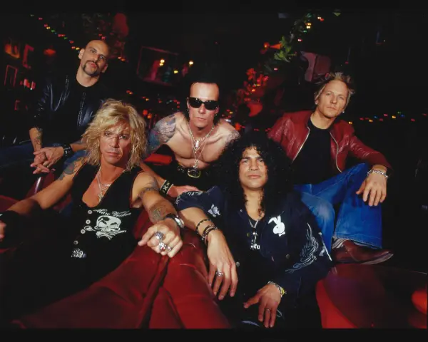 Cinq musiciens, un groupe de slash, posent sur un canapé rouge. Ils portent des vestes en cuir et des bijoux.