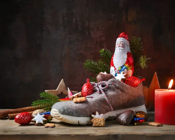 Ein Schuh gefüllt mit Leckereien, daneben ein Schokoladen-Nikolaus, Tannenzweige und eine brennende Kerze.