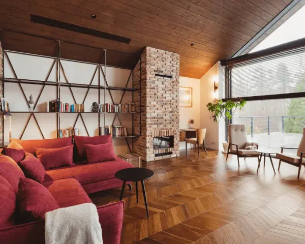 Przytulny salon w stylu wiejskim z czerwoną kanapą, ceglanym kominkiem i regałami pełnymi książek.