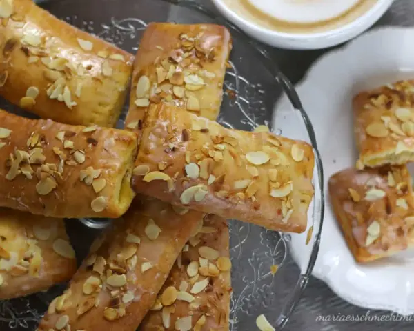 Omas Apfeltaschen Rezept: goldbraune, glasierte Gebäckstücke mit Mandelblättchen, serviert auf einem Glasständer und einem Teller, daneben eine Tasse Kaffee.