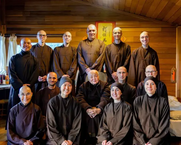 A group of monks and nuns, including an elder monk person in the center, pose for a photo in a room with wooden walls.