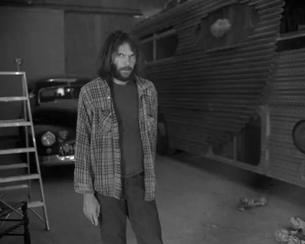 Neil Young, jung und nachdenklich, steht vor einem umgebauten Bus. Eine Leiter und ein Oldtimer im Hintergrund.