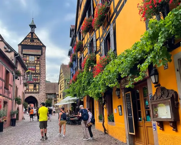 Cobblestone street in a picturesque Alsace town, with colorful half-timbered buildings adorned with flowers. A clock tower stands tall in the background.