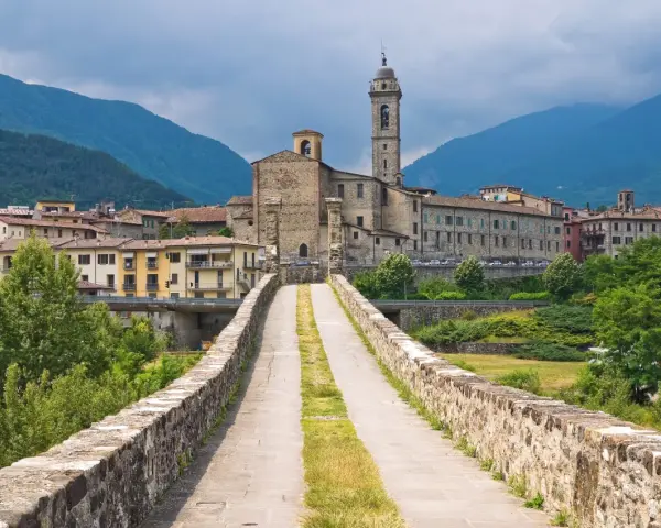 Ponte medievale in pietra che conduce a un borgo con chiesa e campanile, incastonato tra le verdi montagne degli Appennini Emilia Romagna.