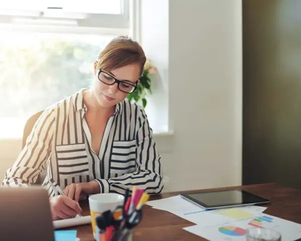 Mujer con gafas trabaja en su escritorio, revisando documentos y gráficos. Quizás está lidiando con gastos de difícil justificación.