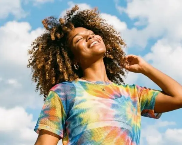 Une femme souriante aux cheveux bouclés, sous un ciel bleu nuageux. Elle porte un t-shirt tie-dye coloré. Une belle image pour apprendre comment rendre une photo flou.