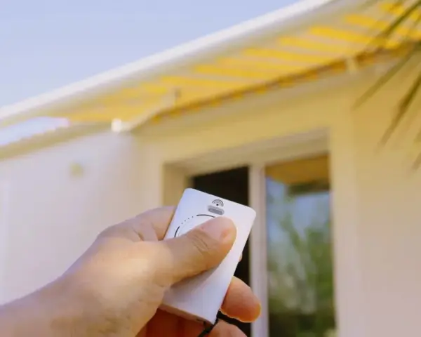 Main tenant une télécommande pour le réglage store banne manuel. Le store jaune est déployé sur une maison blanche.