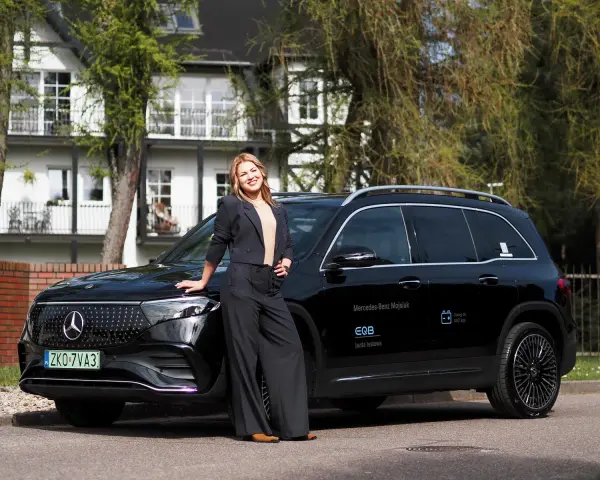 Kobieta w czarnym garniturze opiera się o czarnego Mercedesa EQB na tle białego domu.