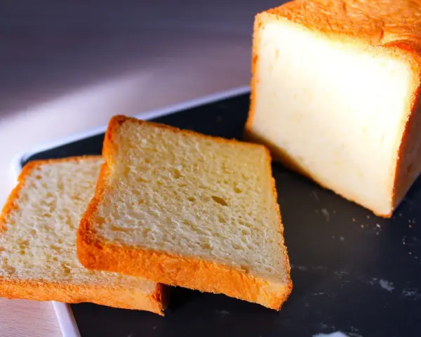 Pain de mie moelleux, parfait pour une recette pain de mie boulangerie maison. Deux tranches dorées et un pain entier sur un plateau sombre.