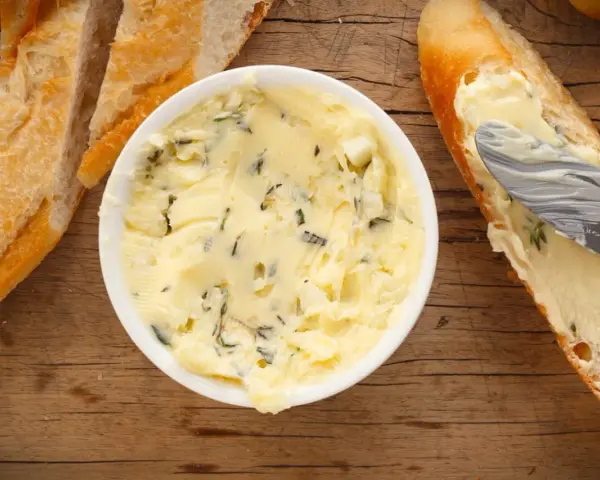 Knoblauchbutter selber machen ist einfach! Hier ist eine Schüssel mit selbstgemachter Knoblauchbutter, daneben Baguettescheiben und ein Messer, das Butter auf ein Brot streicht.
