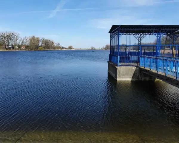 Spokojne jezioro średzkie z błękitną budowlą i pomostem. W tle drzewa i zabudowania.