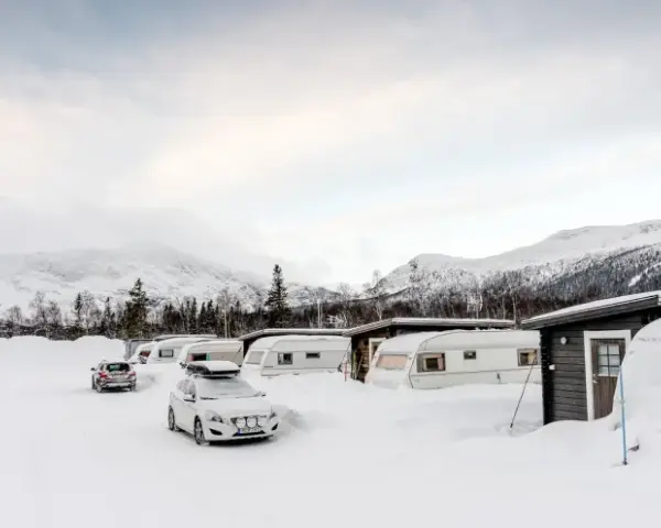 Vinterlandskap vid moskosel camping med snötäckta husvagnar och bilar, berg i bakgrunden.