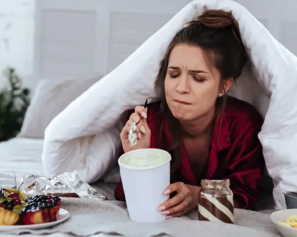 Ragazza sotto le coperte mangia gelato, dolci e pasta. La sua espressione suggerisce una forte fame pre ciclo.