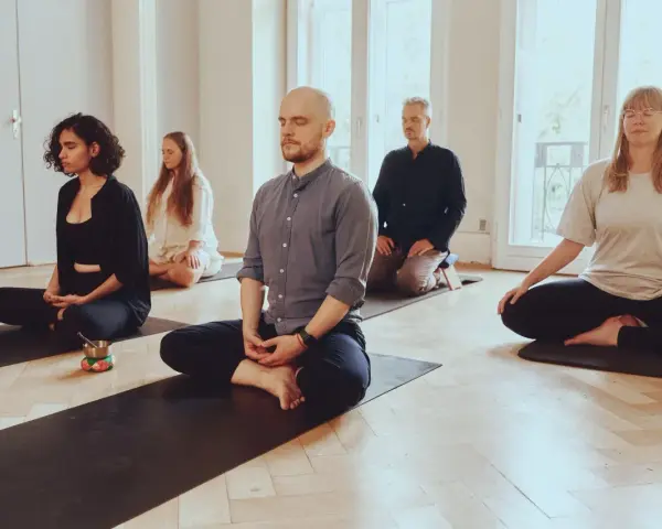 Menschen in einem hellen Raum meditieren auf Matten. Die Meditation wirkt beruhigend und fördert die Konzentration.